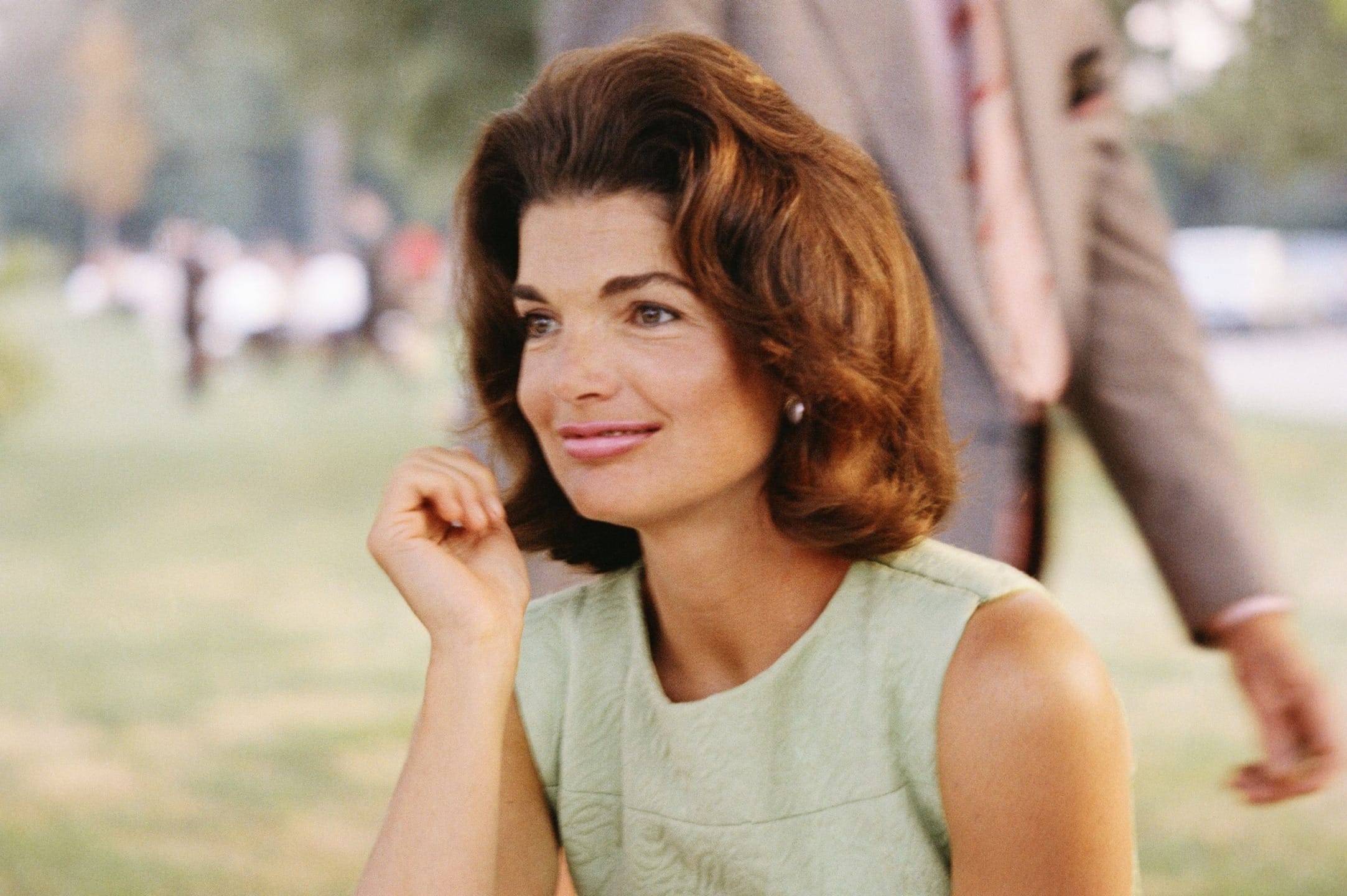 Woman in a green dress sitting outdoors, smiling with a blurred person in the background.