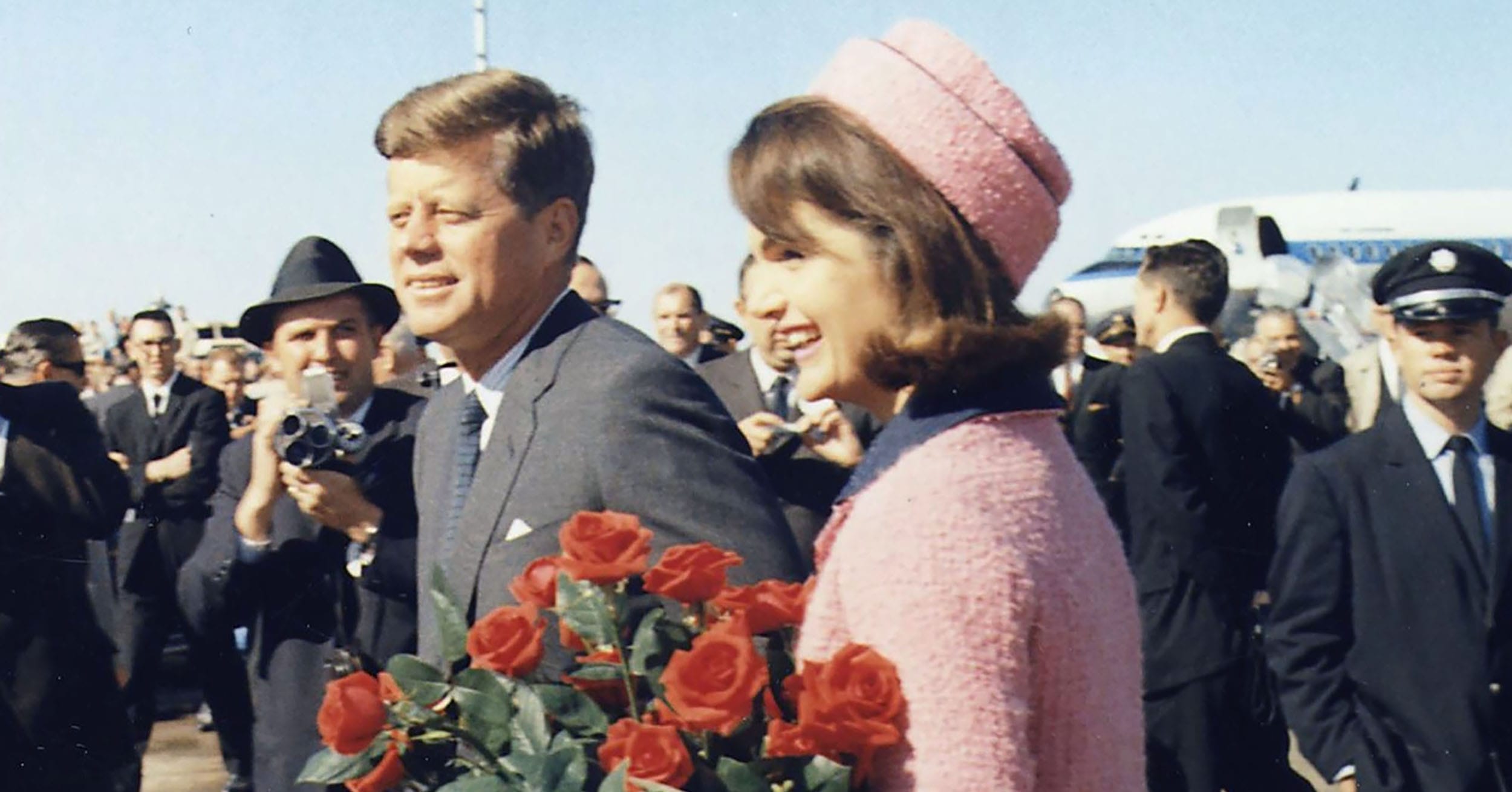 A man and woman walking outside surrounded by people, with the woman holding a bouquet of red roses.