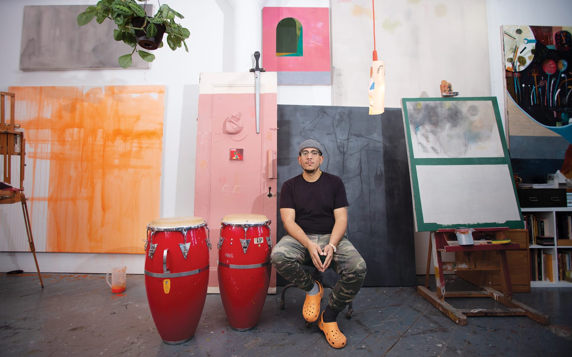 Artist sitting in a studio with paintings, red conga drums, and wearing camo pants and orange shoes.