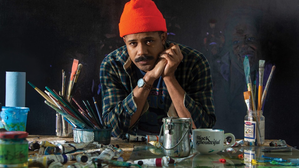 Artist wearing a red beanie, surrounded by painting supplies, leaning on a cluttered table with paint tubes and brushes.