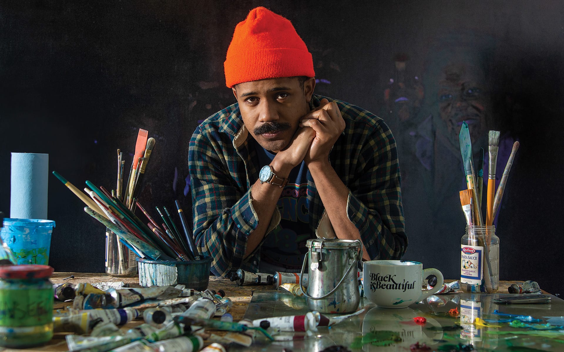 Artist wearing a red beanie, surrounded by painting supplies, leaning on a cluttered table with paint tubes and brushes.