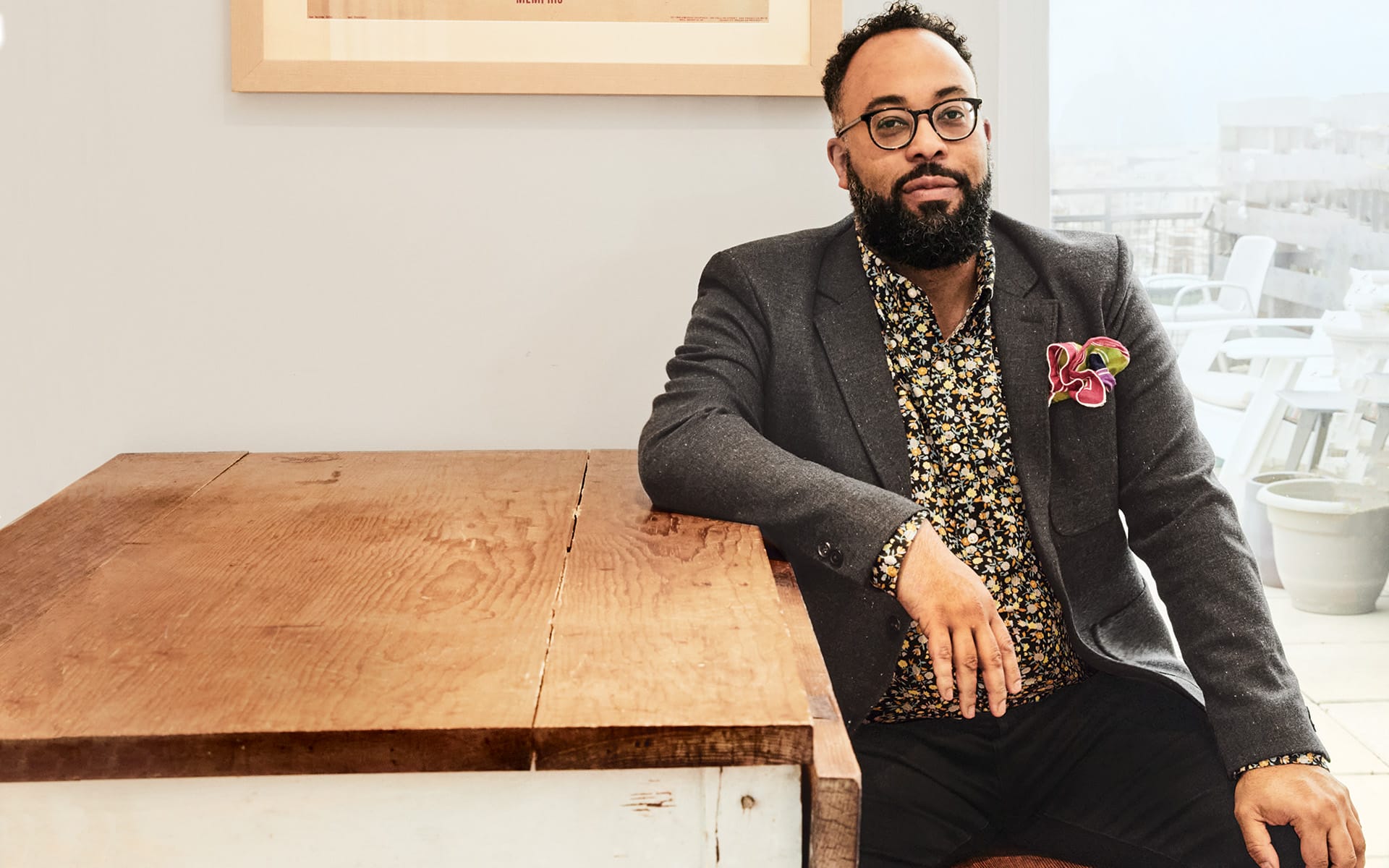 Man sitting at a wooden table wearing glasses and a patterned shirt under a dark blazer with a colorful pocket square.