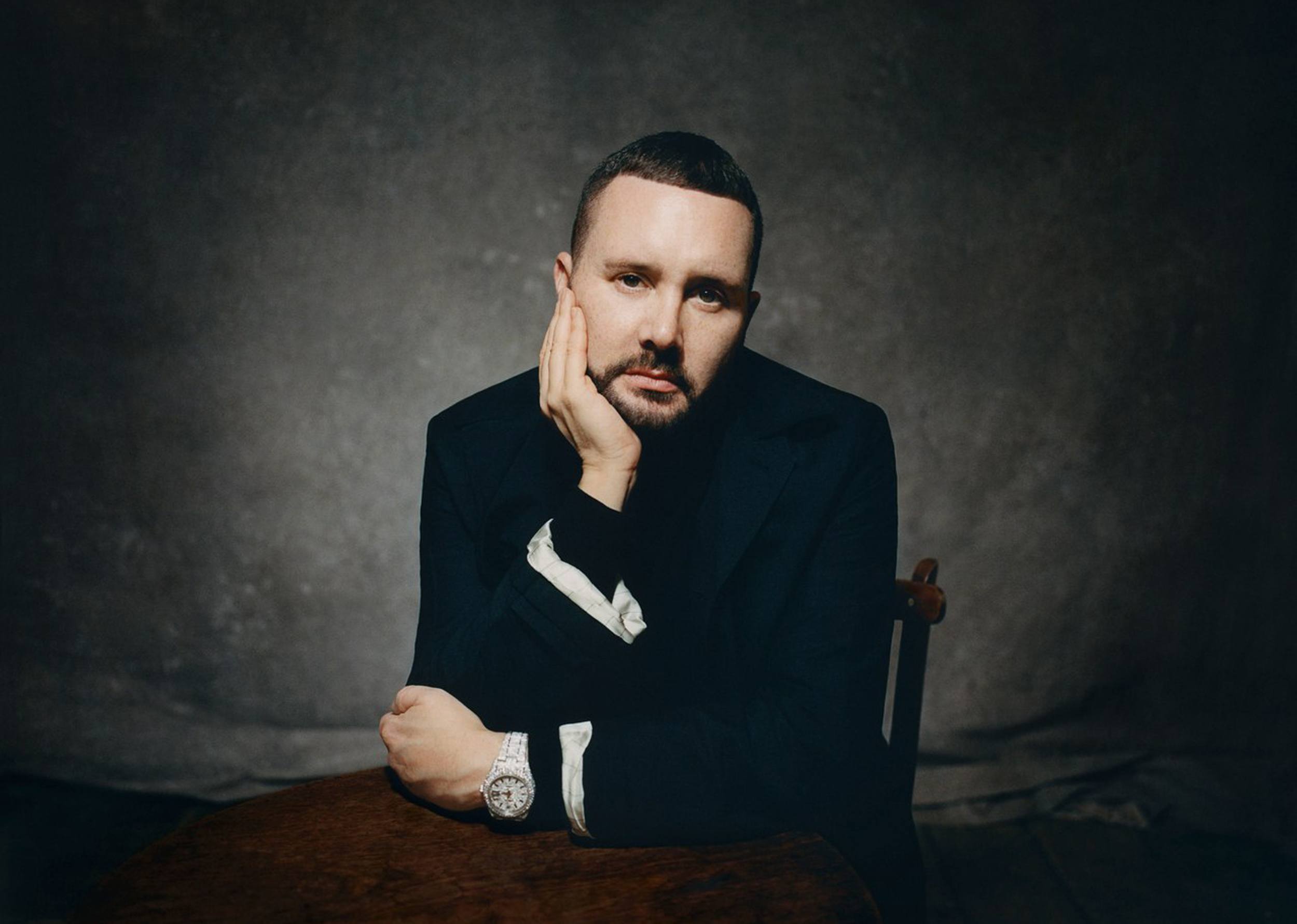 Person sitting at a table, resting their face on one hand, wearing a dark suit with a watch, against a dark backdrop.