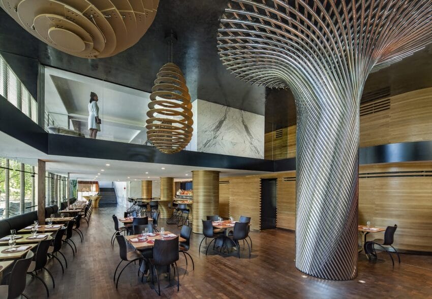 Modern restaurant interior with unique spiral columns, wooden floors, and elegant table settings. Upper level with person visible.