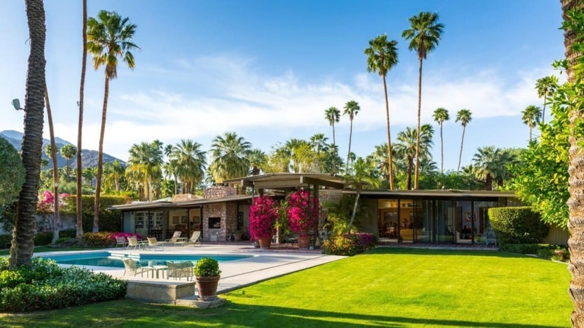 Modern home with large windows, pool, and palm trees in a lush backyard setting with a mountain in the background.