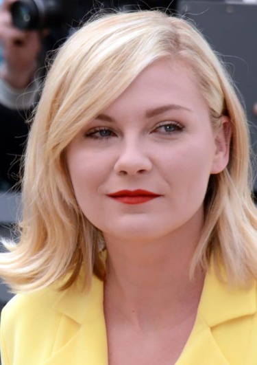 Person with short blonde hair wearing a yellow jacket, looking ahead with a neutral expression in a candid outdoor setting.