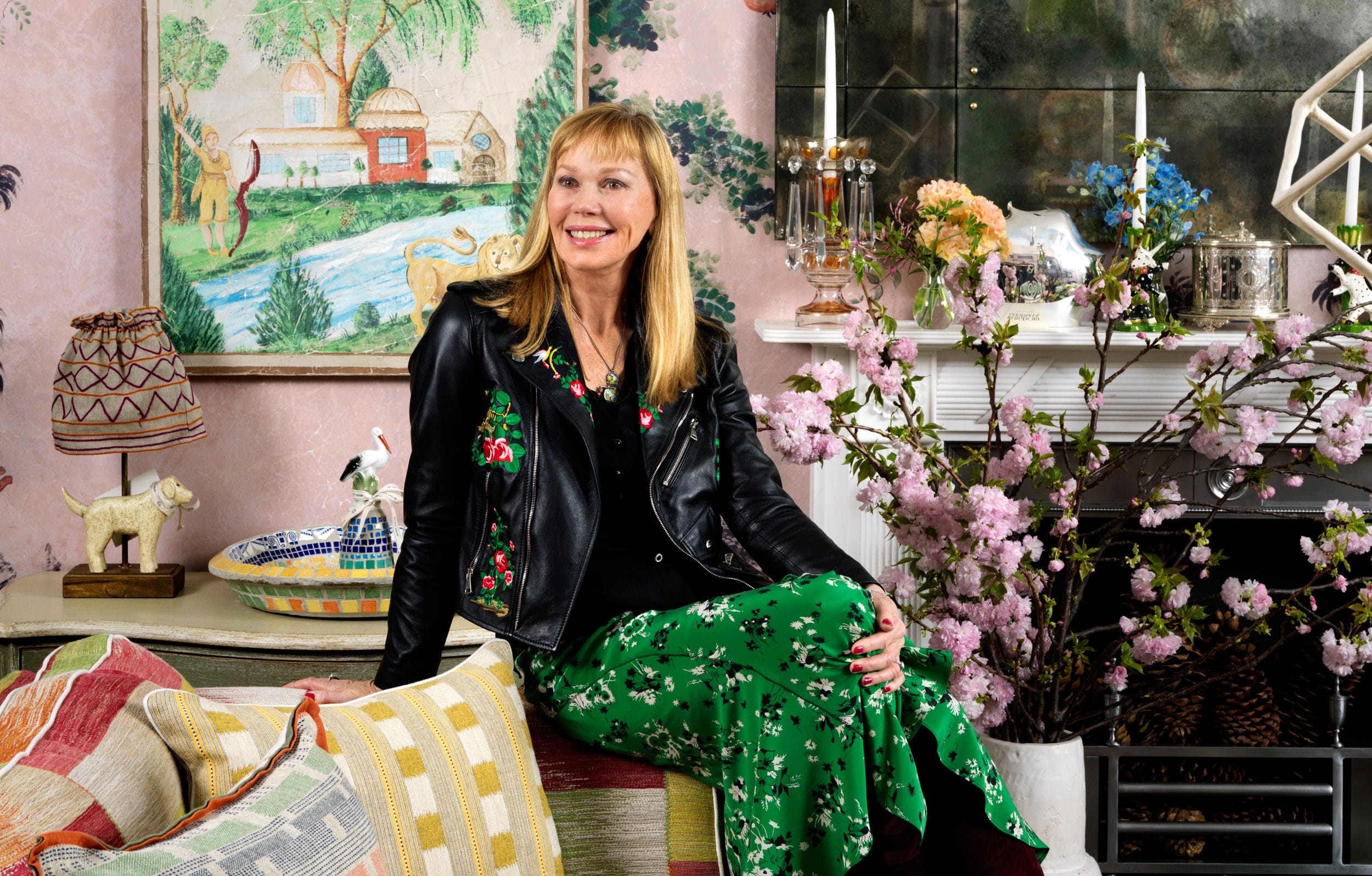 Person in a floral outfit sitting in a colorful, eclectic room with art, plants, and decorative objects in the background.