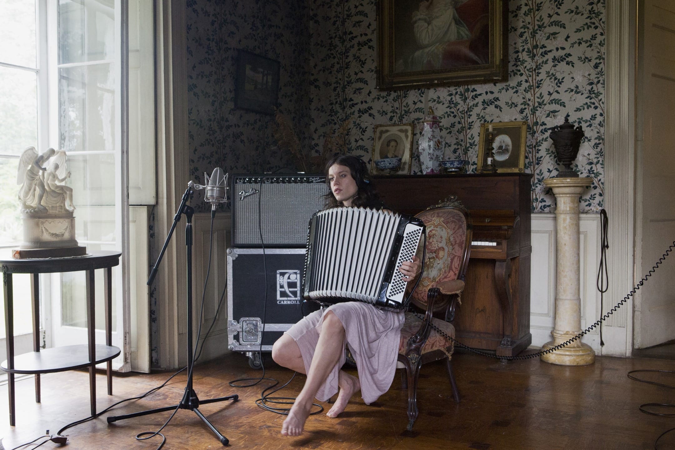 A person sitting barefoot in a vintage room, playing an accordion near a microphone and a guitar amplifier.