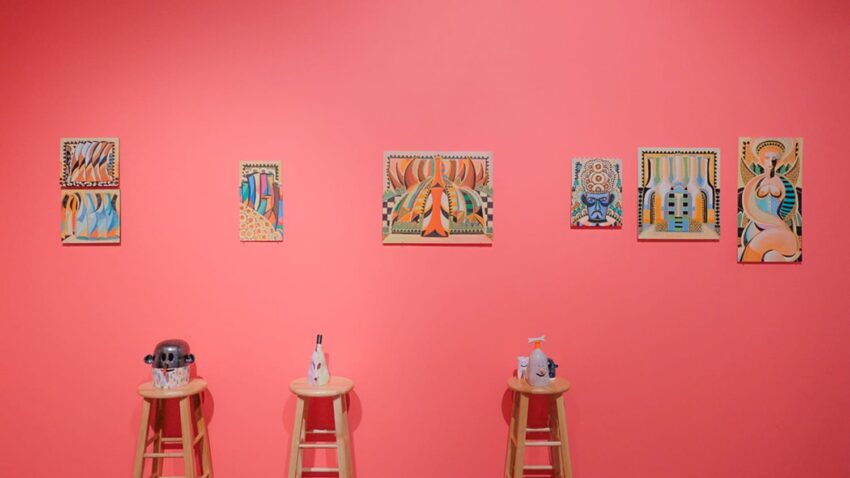 Abstract art pieces on a pink wall above three stools with various items on them in a gallery setting.