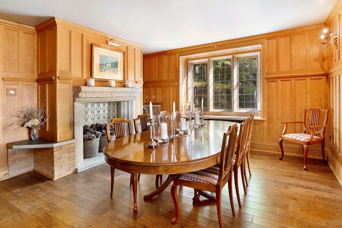 Elegant dining room with wooden panel walls, large wooden table, chairs, candles, fireplace, and large window with natural light.