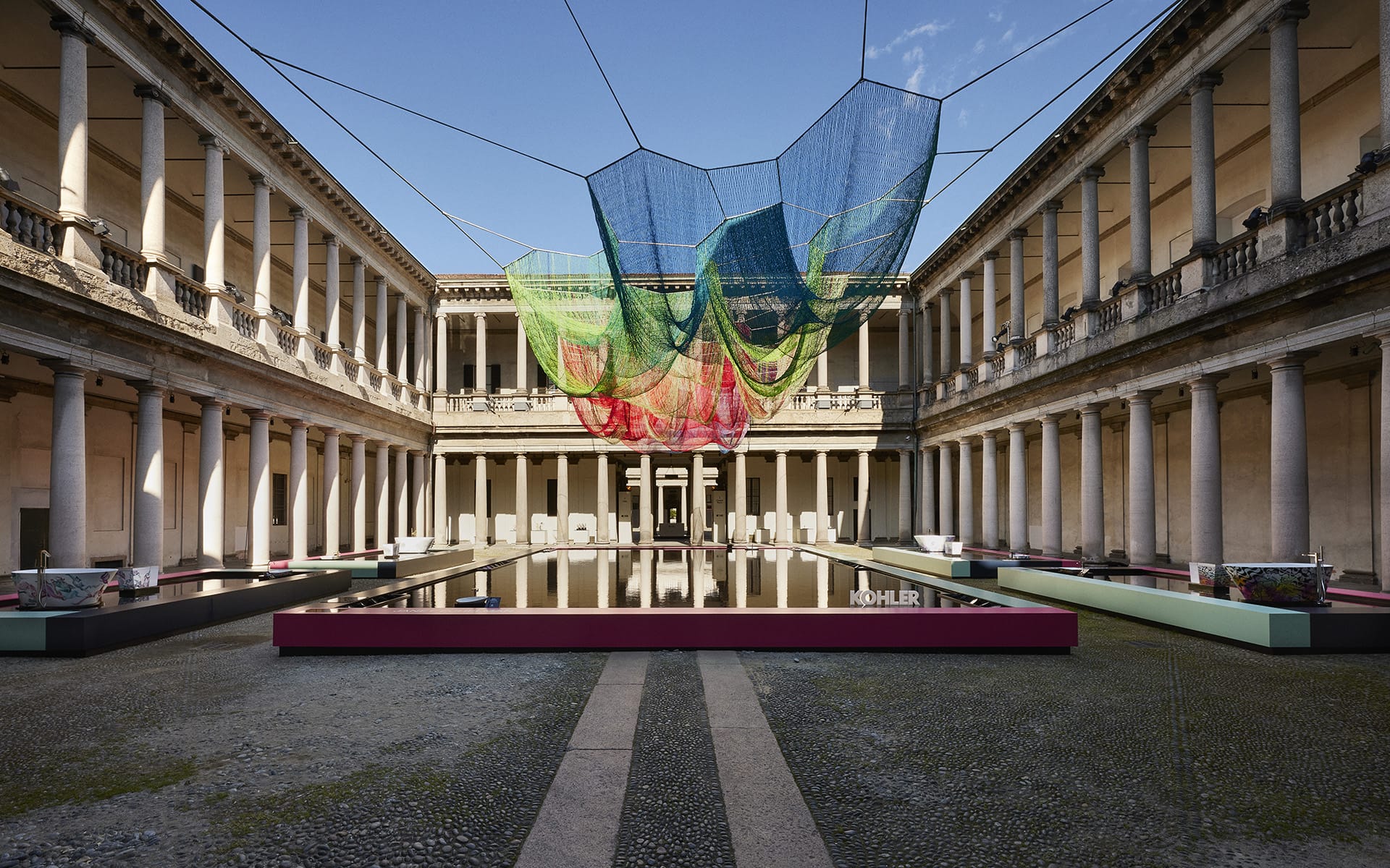 Colorful net-like art installation suspended above a reflective water surface in a classical courtyard with columns.