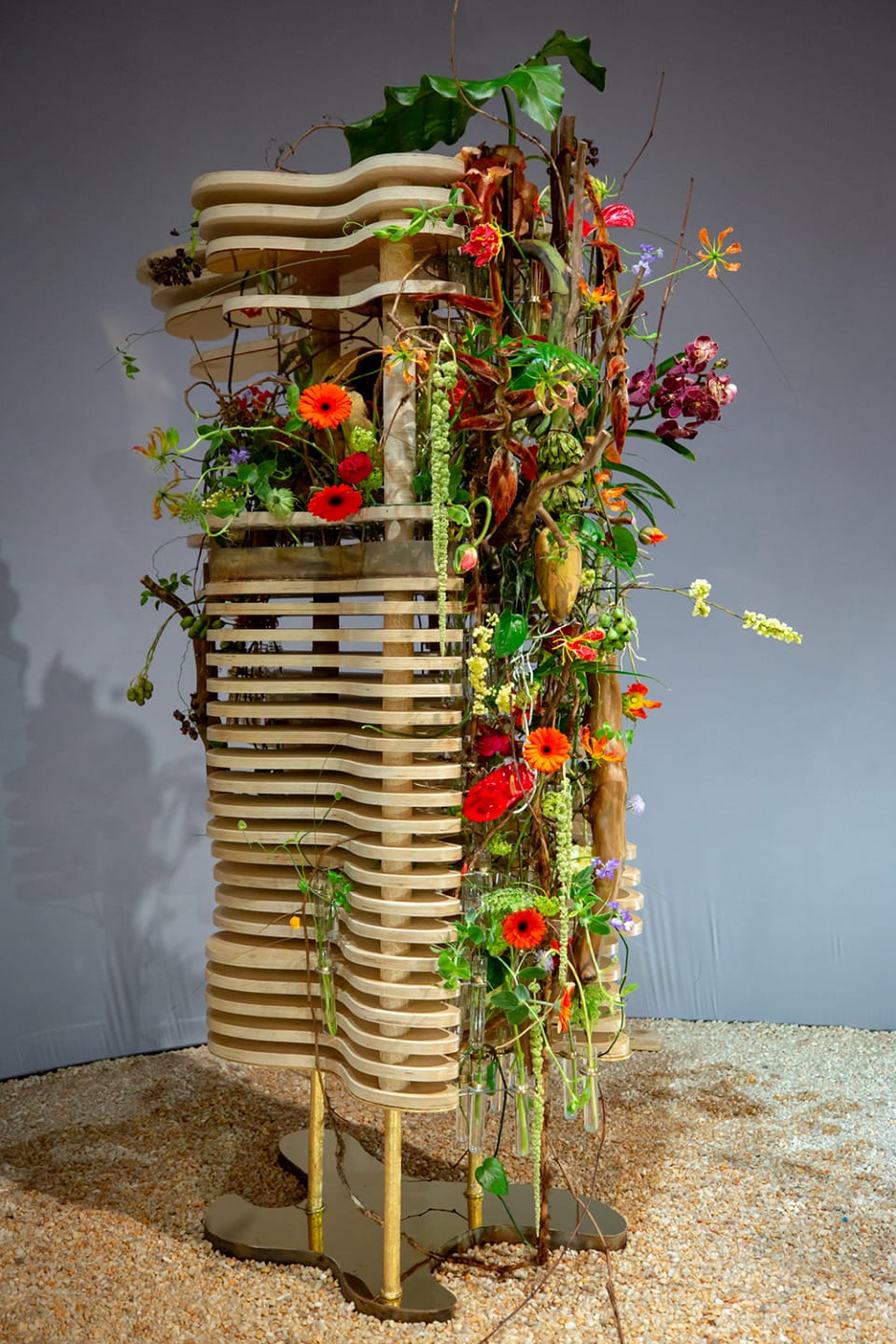 Abstract wooden sculpture adorned with colorful flowers and greenery displayed against a neutral background.