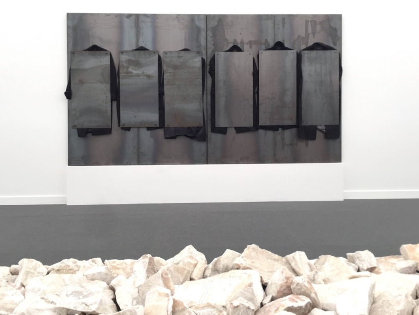 Metallic abstract art on a white wall above a bed of white stones in a modern gallery setting.