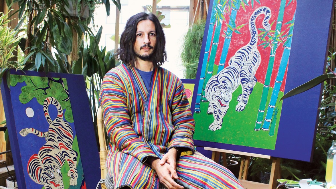 Artist in striped robe seated between two colorful tiger paintings in a studio filled with greenery.