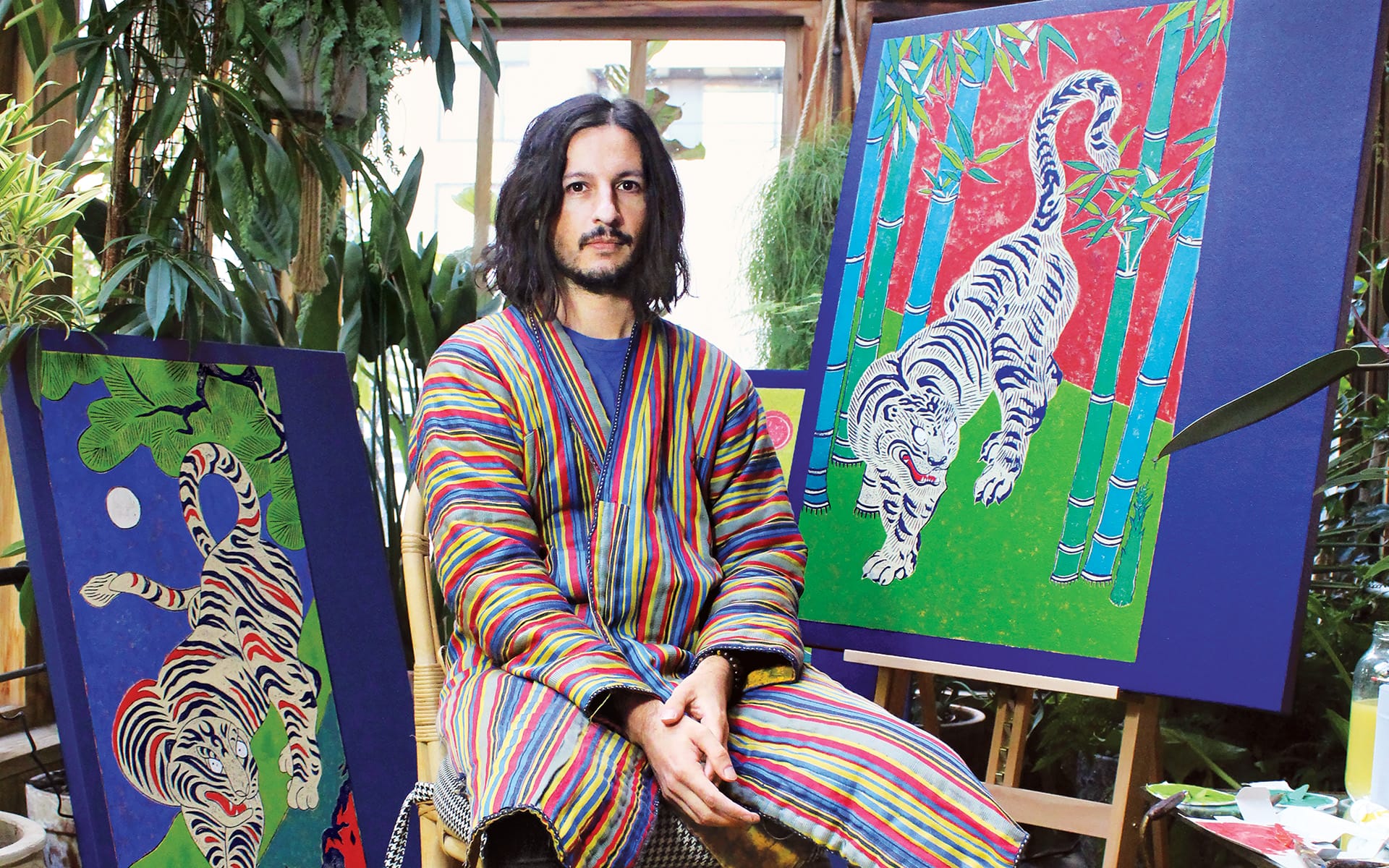 Artist in striped robe seated between two colorful tiger paintings in a studio filled with greenery.