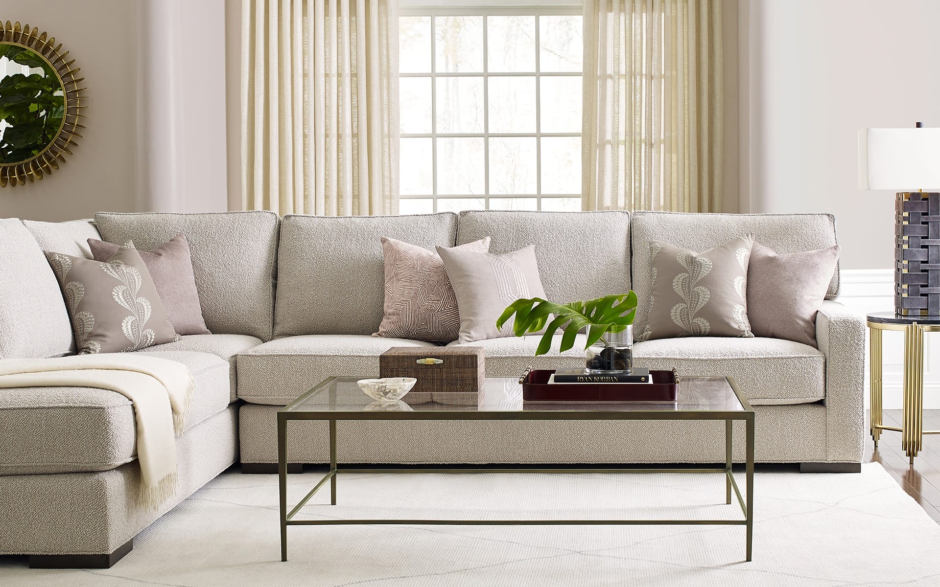 Modern living room with a gray sectional sofa, decorative pillows, a glass coffee table, and a window with sheer curtains.