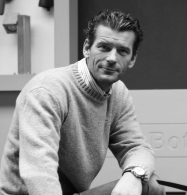 Man in a sweater and striped shirt sitting indoors, looking at the camera with a relaxed pose, wearing a watch.