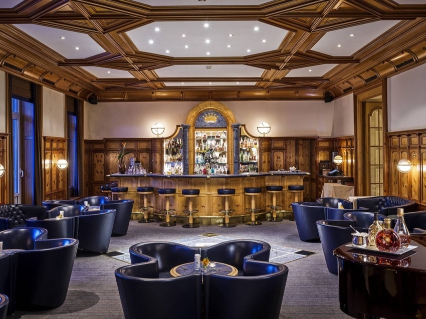 Elegant bar with wooden decor, plush navy seating, and a well-stocked backlit bar under a coffered ceiling.