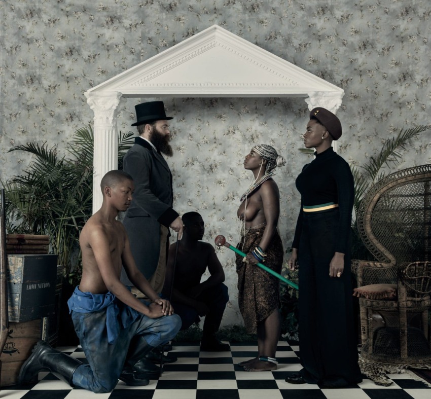 A group of people dressed in diverse, culturally inspired clothing, posed in an ornate, staged setting with checkered floor.