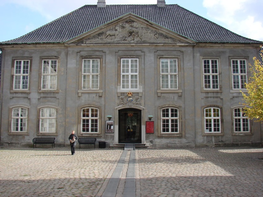 Historic European building facade with ornate details, symmetrical windows, and a cobblestone courtyard on a sunny day.