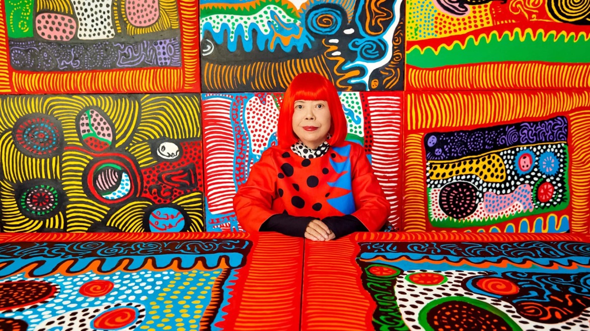 Person in a vibrant red outfit sitting against a colorful, patterned artistic background featuring swirls and dots.