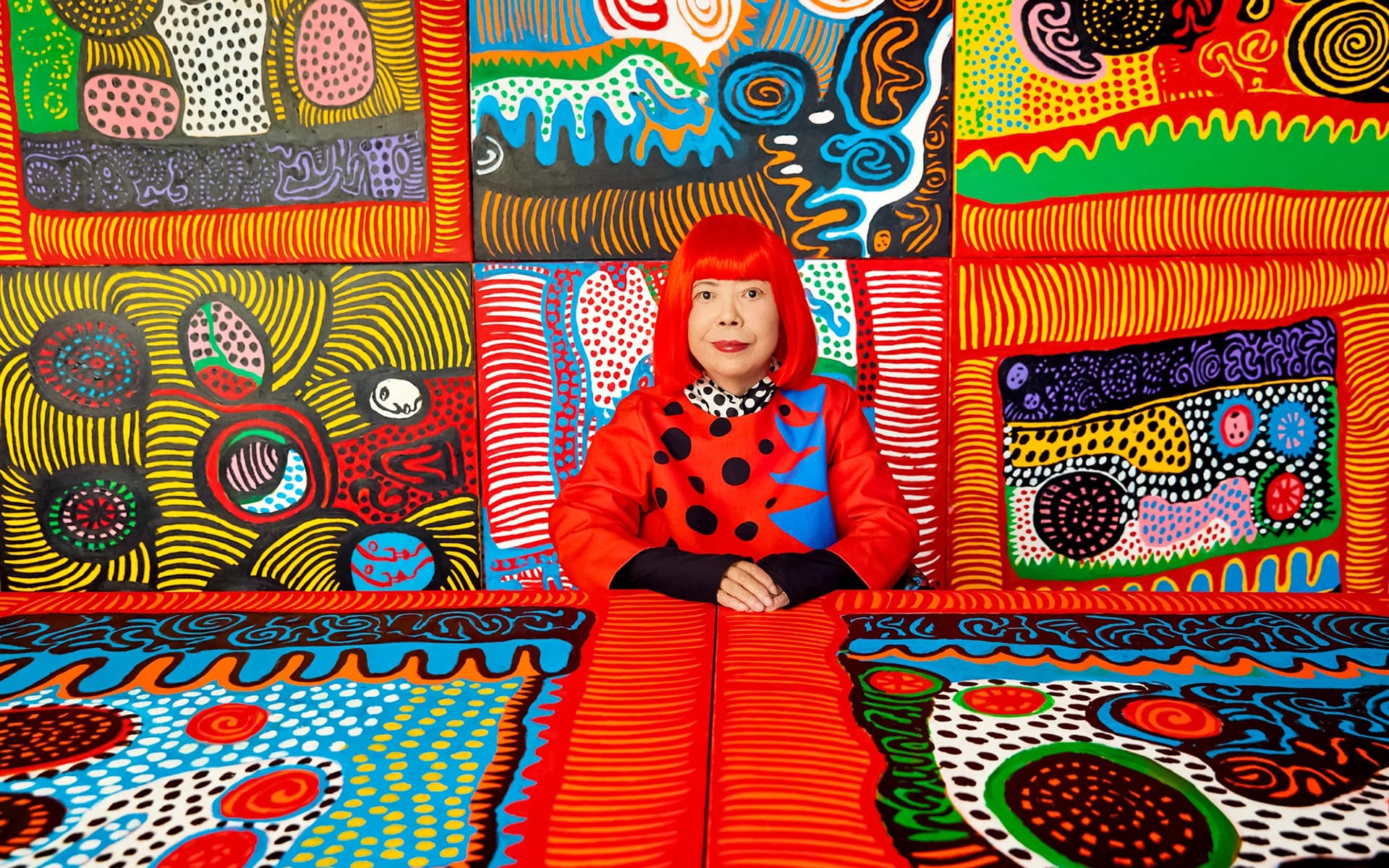 Person in a vibrant red outfit sitting against a colorful, patterned artistic background featuring swirls and dots.
