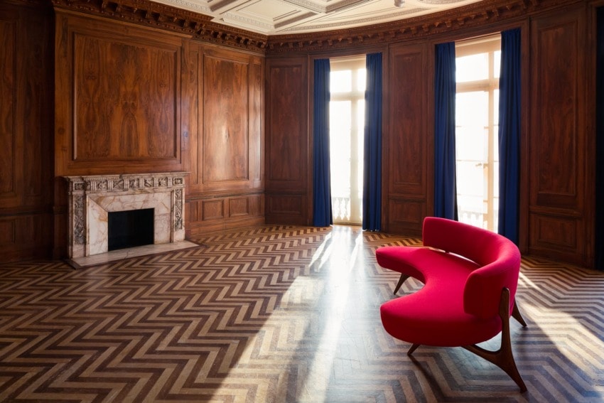Red sofa in an elegant room with wooden walls, chevron-patterned floor, and large windows with blue curtains.