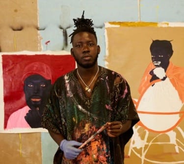Artist standing in front of colorful paintings, holding a paintbrush, wearing a paint-splattered shirt.