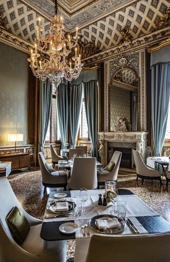 Elegant dining room with ornate chandelier, high ceilings, luxurious decor, set tables, and large windows with drapes.