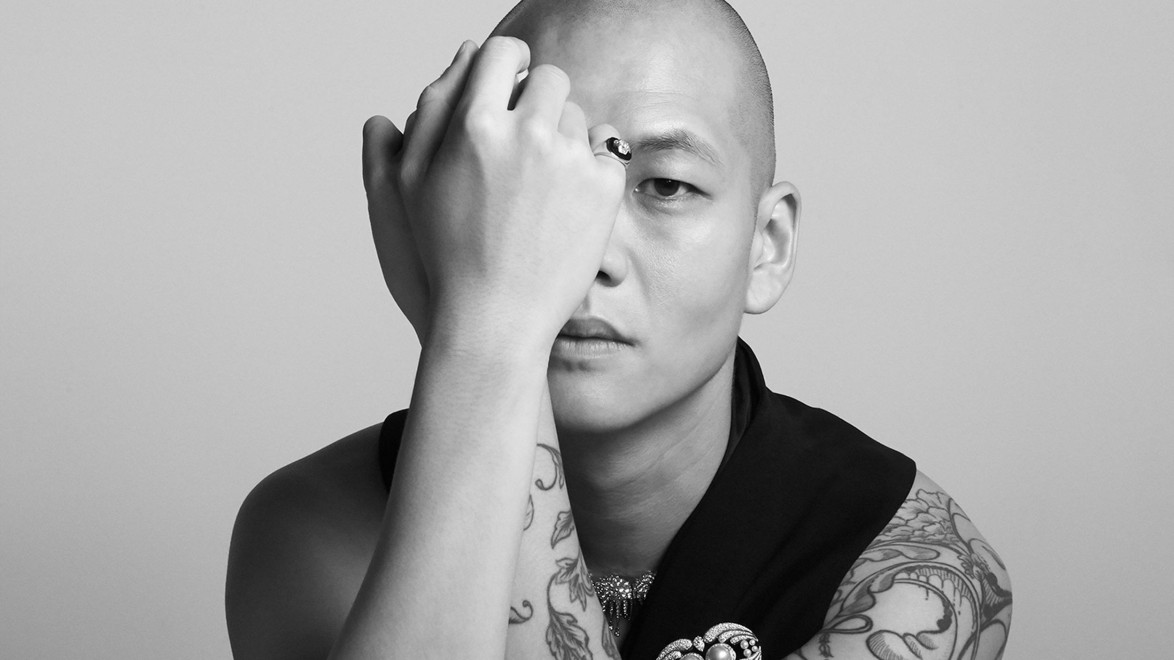 Person with shaved head and tattoos covering eye with hand, wearing a dark garment and decorative brooch, neutral backdrop.