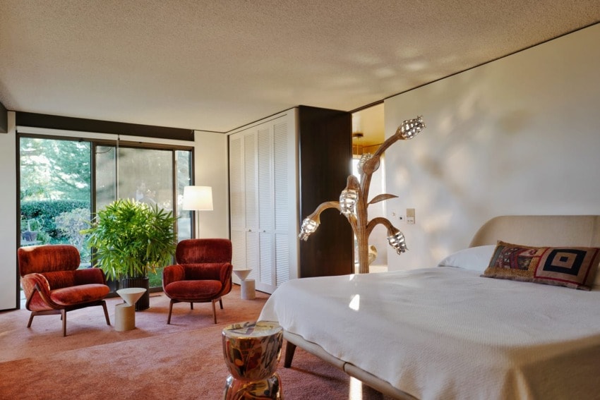 Mid-century modern bedroom with vintage red chairs, unique lamp, and sliding glass doors leading to a lush outdoor view.