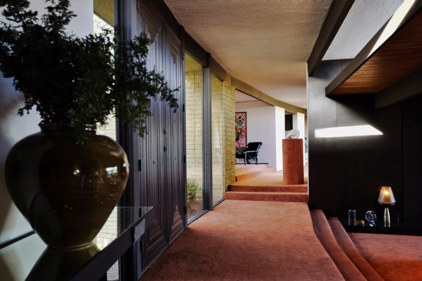 Mid-century modern interior with large windows, brick walls, and a plant in the foreground, showcasing vintage decor elements.