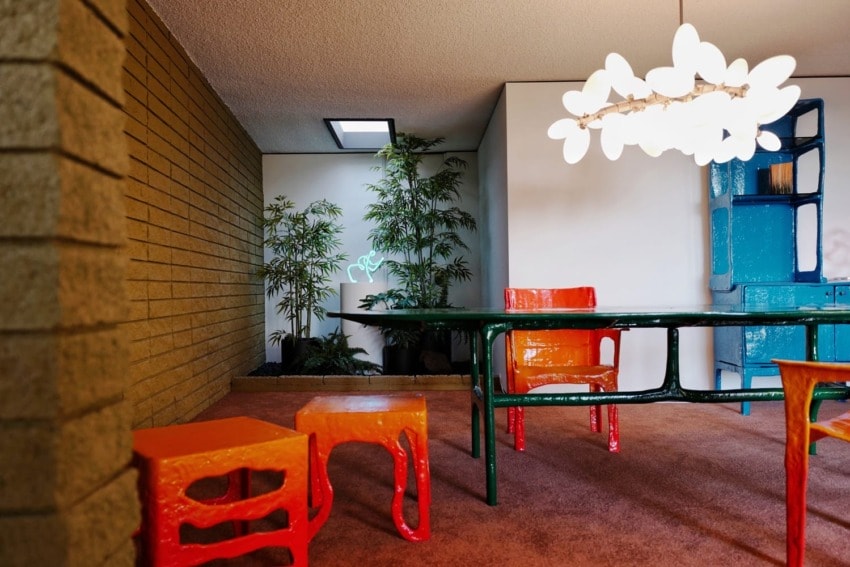 Modern room with colorful chairs, green table, white chandelier, indoor plants, and natural lighting from a skylight.