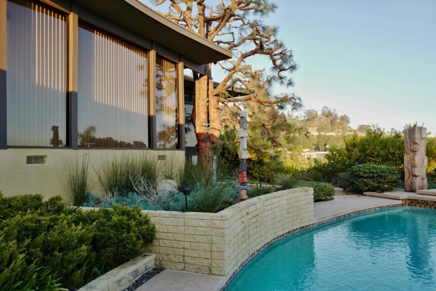 Modern house with large windows, surrounded by lush greenery and a clear blue swimming pool on a sunny day.