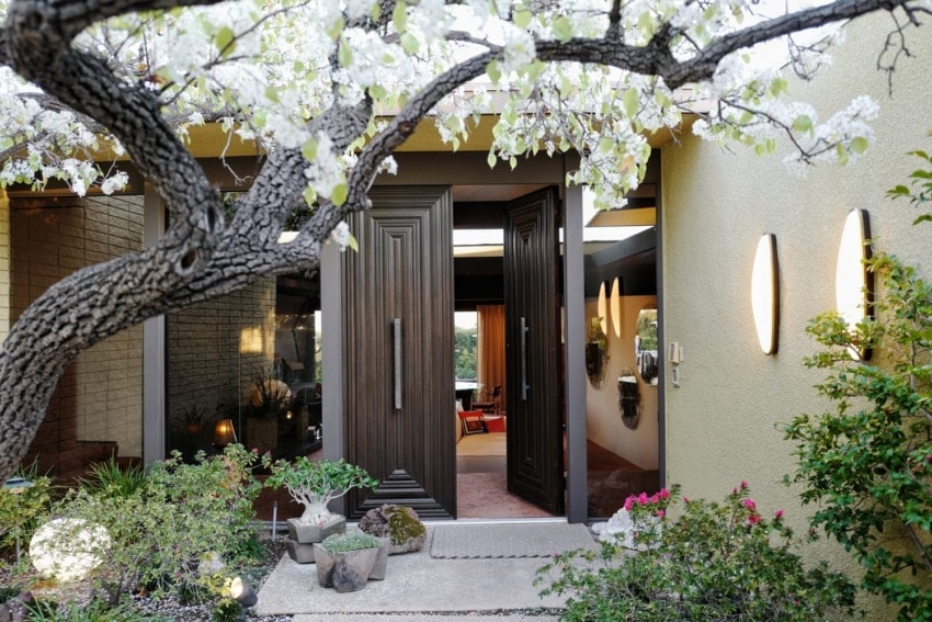Modern house entrance with wooden double doors, surrounded by green plants and white flowering tree branches.