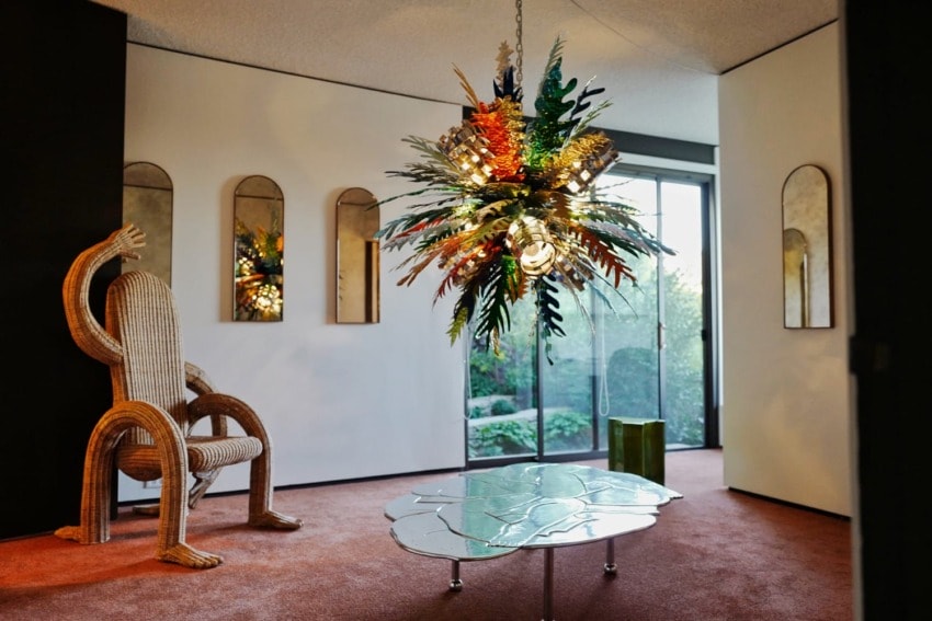 Eclectic modern living room with vibrant abstract chandelier, mirrored wall, artistic chair, and glass doors opening to a garden.