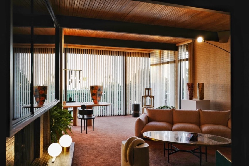 Mid-century modern living room with large windows, a curved sofa, round marble table, and floor-to-ceiling blinds.