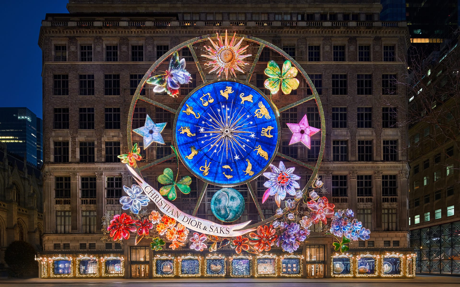 Festive light display on a building facade with zodiac wheel and floral decorations at night.