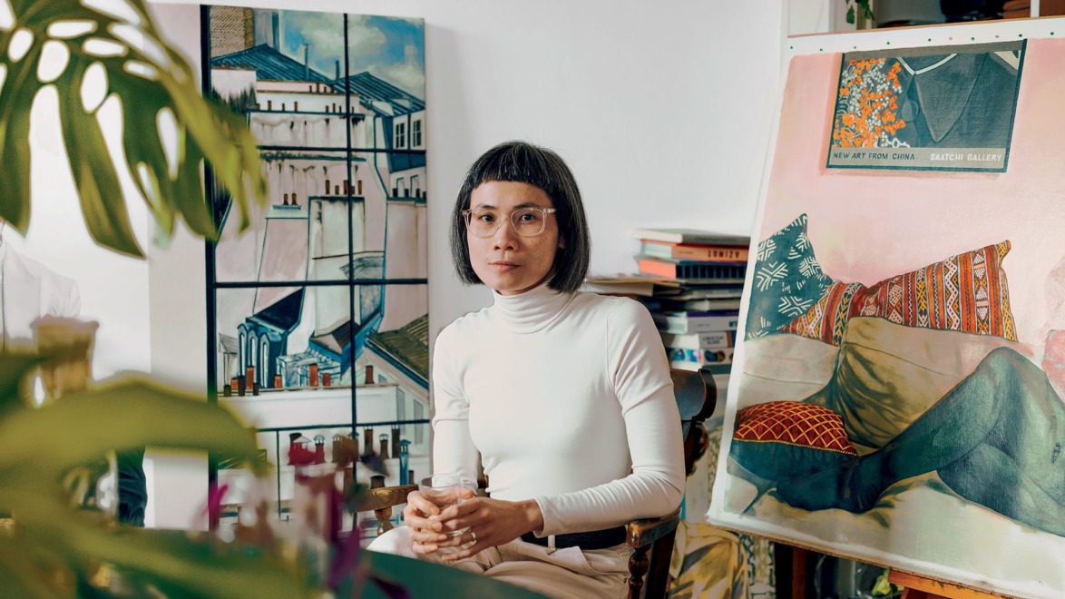 Artist seated in studio with paintings around, holding a glass and wearing a white turtleneck, surrounded by canvases.