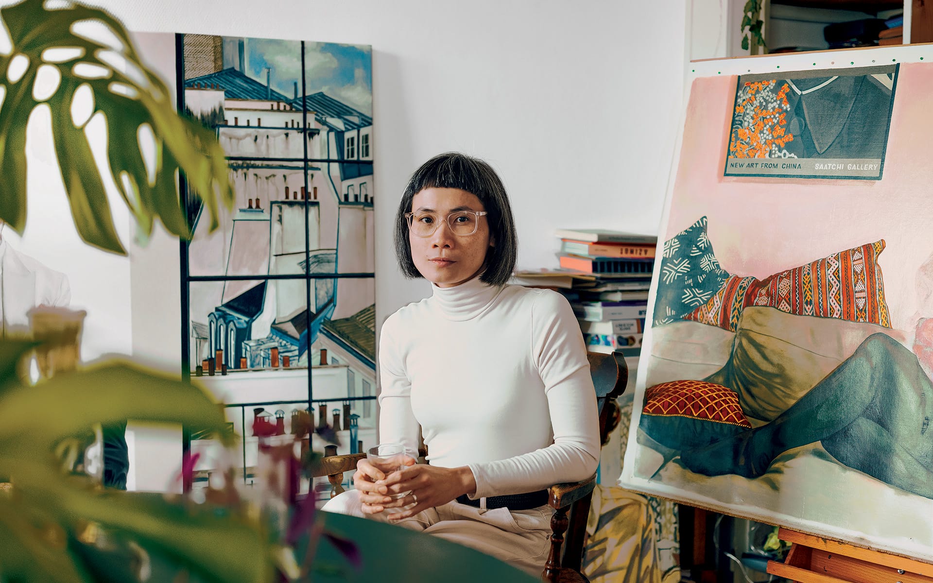Artist seated in studio with paintings around, holding a glass and wearing a white turtleneck, surrounded by canvases.