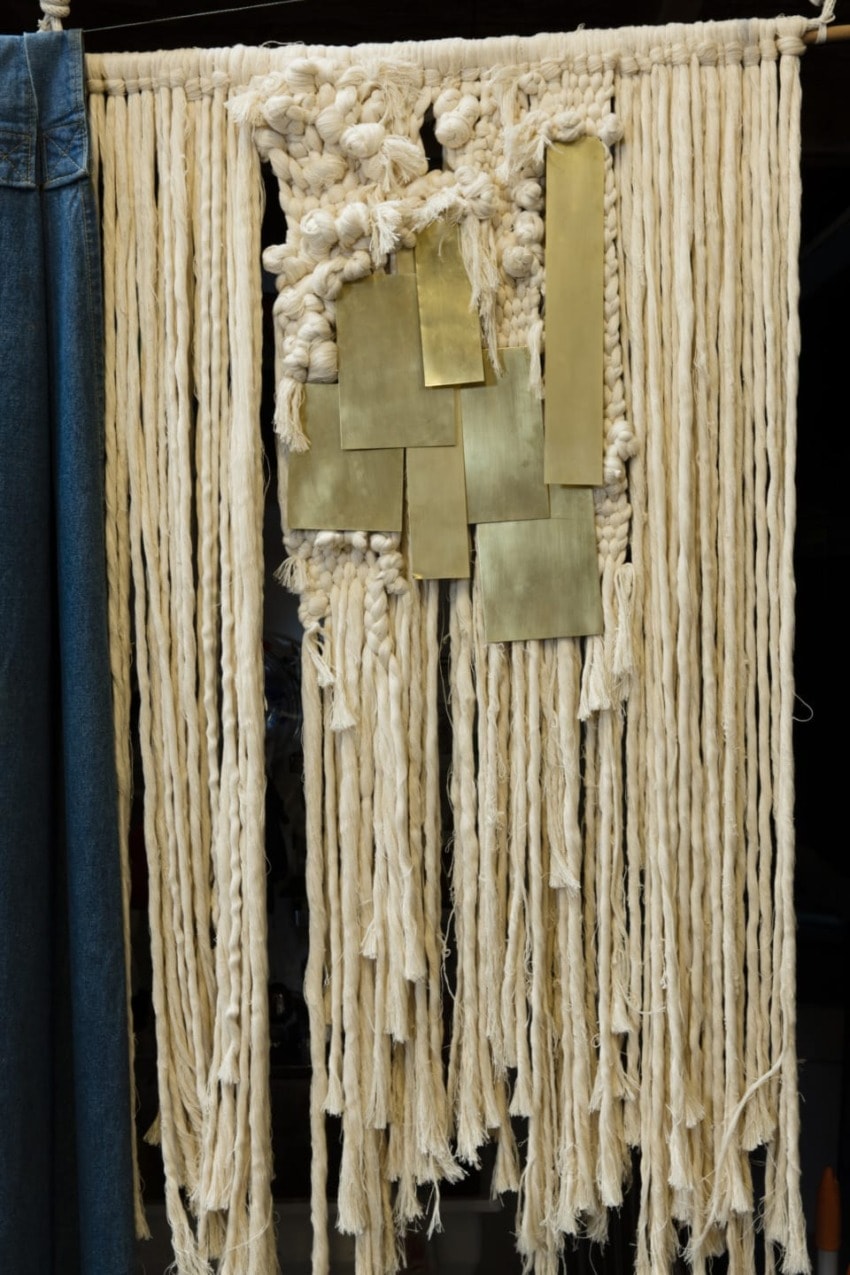 Handwoven macramé wall hanging with long fringe and gold metal accents, displayed against a denim fabric background.