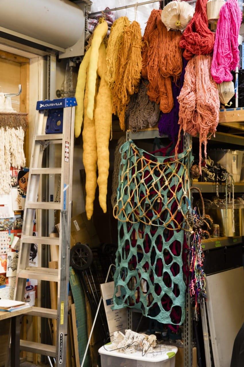 Art studio with colorful yarns hanging, ladder in the foreground, and a large net-like piece draped on the wall.