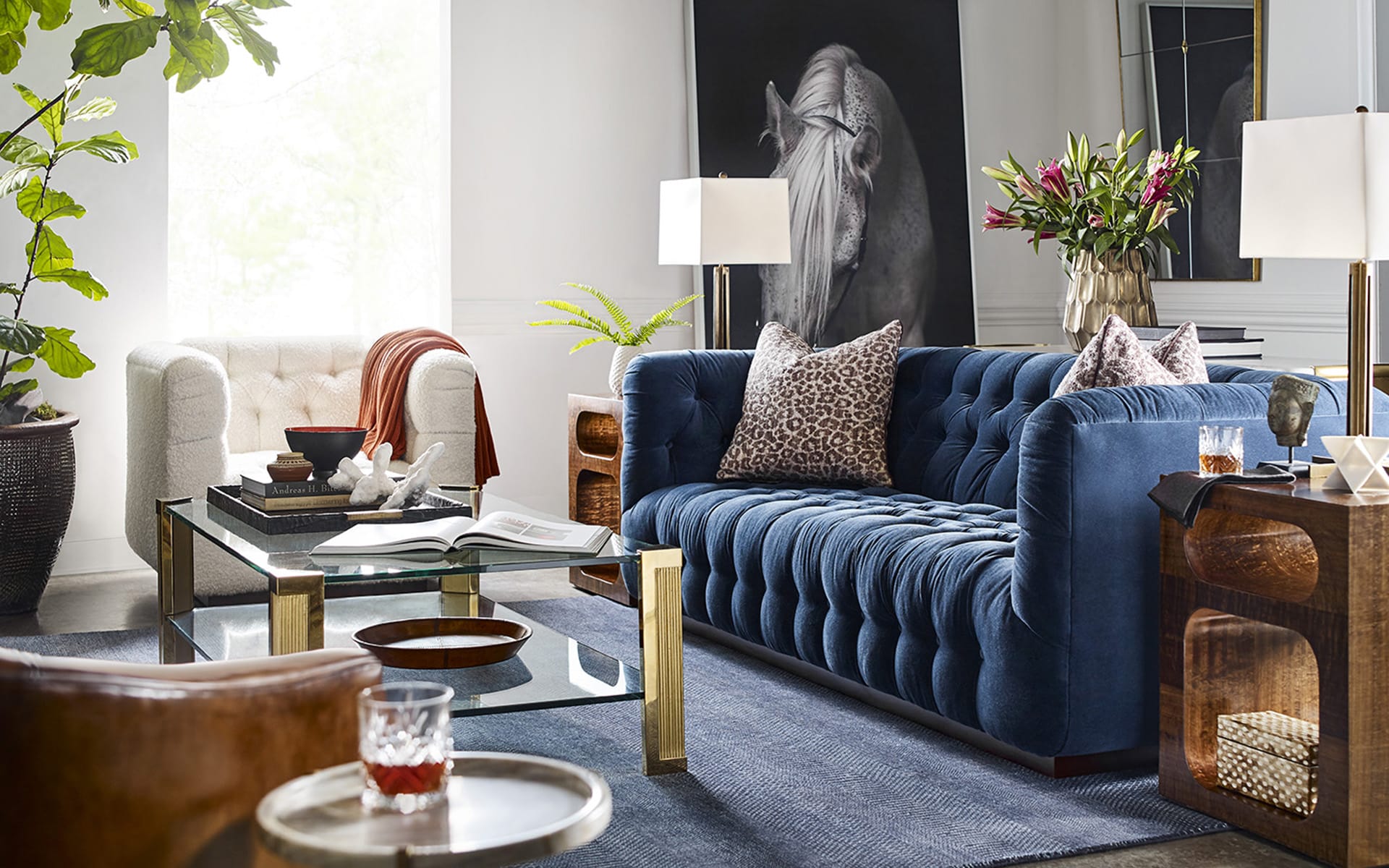 Modern living room with a blue velvet sofa, glass coffee table, and decorative plants and artwork.