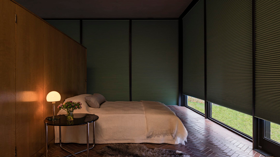 Cozy modern bedroom with a large window, a neatly made bed, soft lighting, a side table with flowers, and a rug on brick flooring.