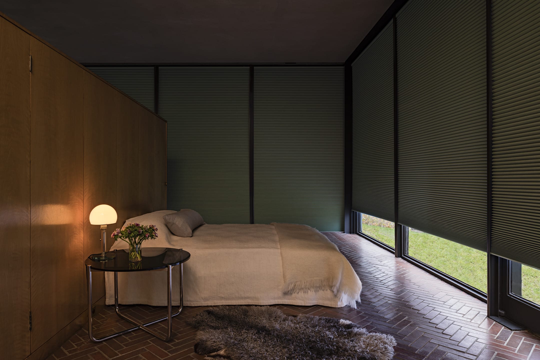 Cozy modern bedroom with a large window, a neatly made bed, soft lighting, a side table with flowers, and a rug on brick flooring.