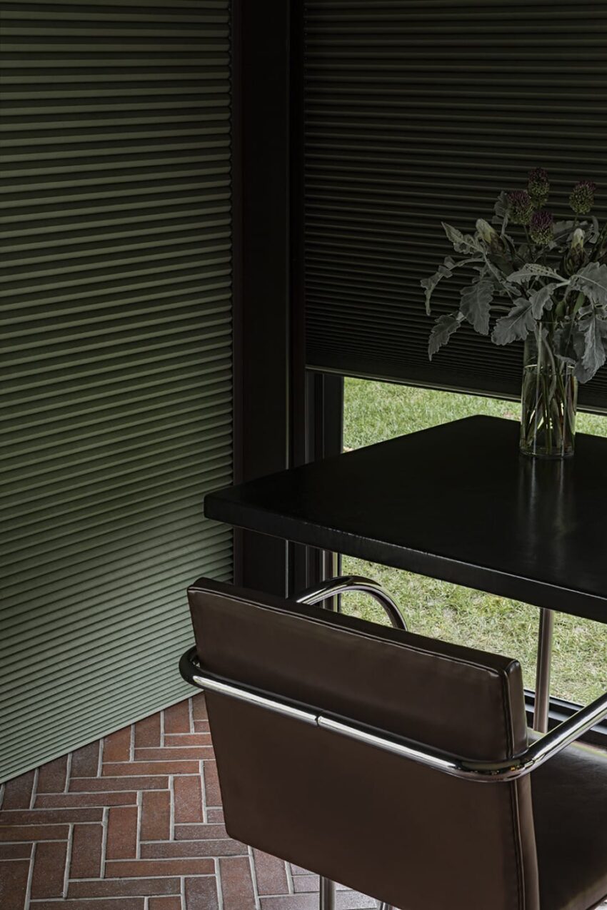 Dark modern dining area with a black table, brown chair, and plant arrangement, featuring green blinds and brick flooring.