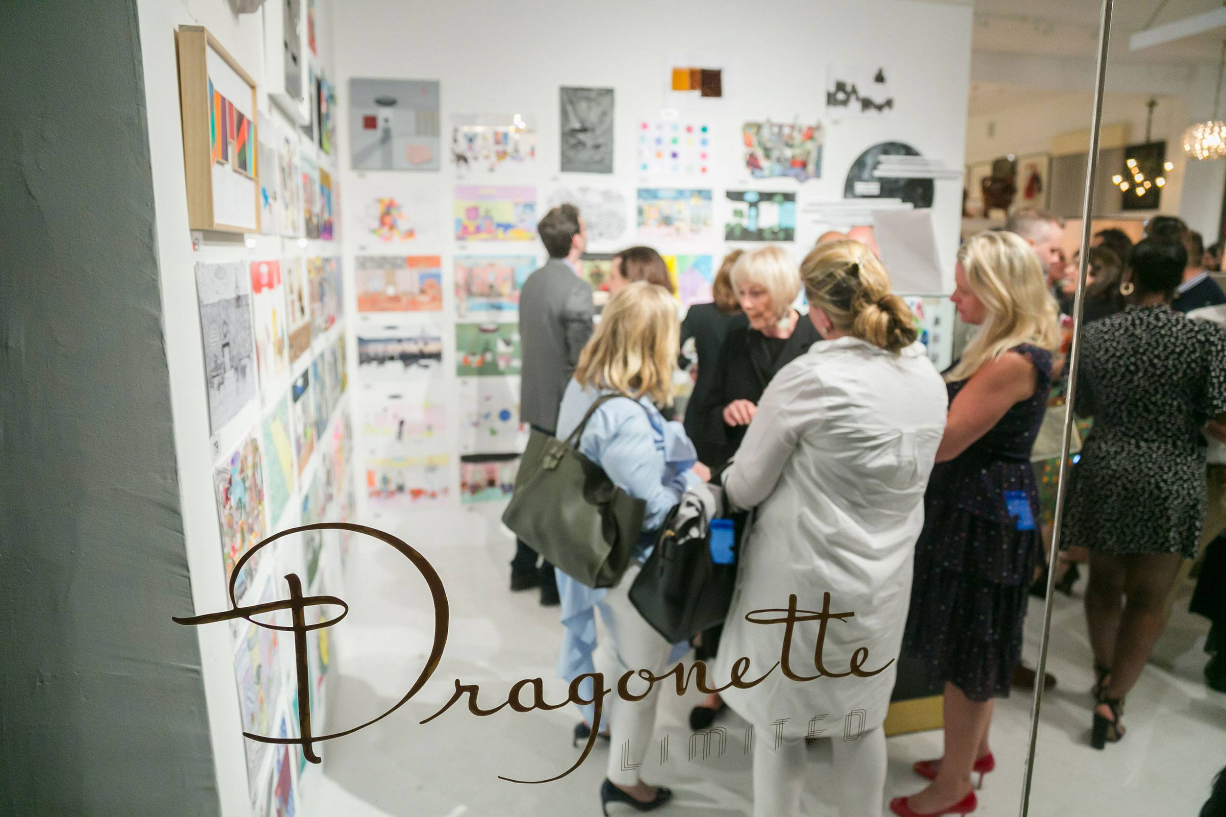 People gathered in an art gallery, viewing colorful artworks displayed on the walls, with "Dragonette" written on a glass door.