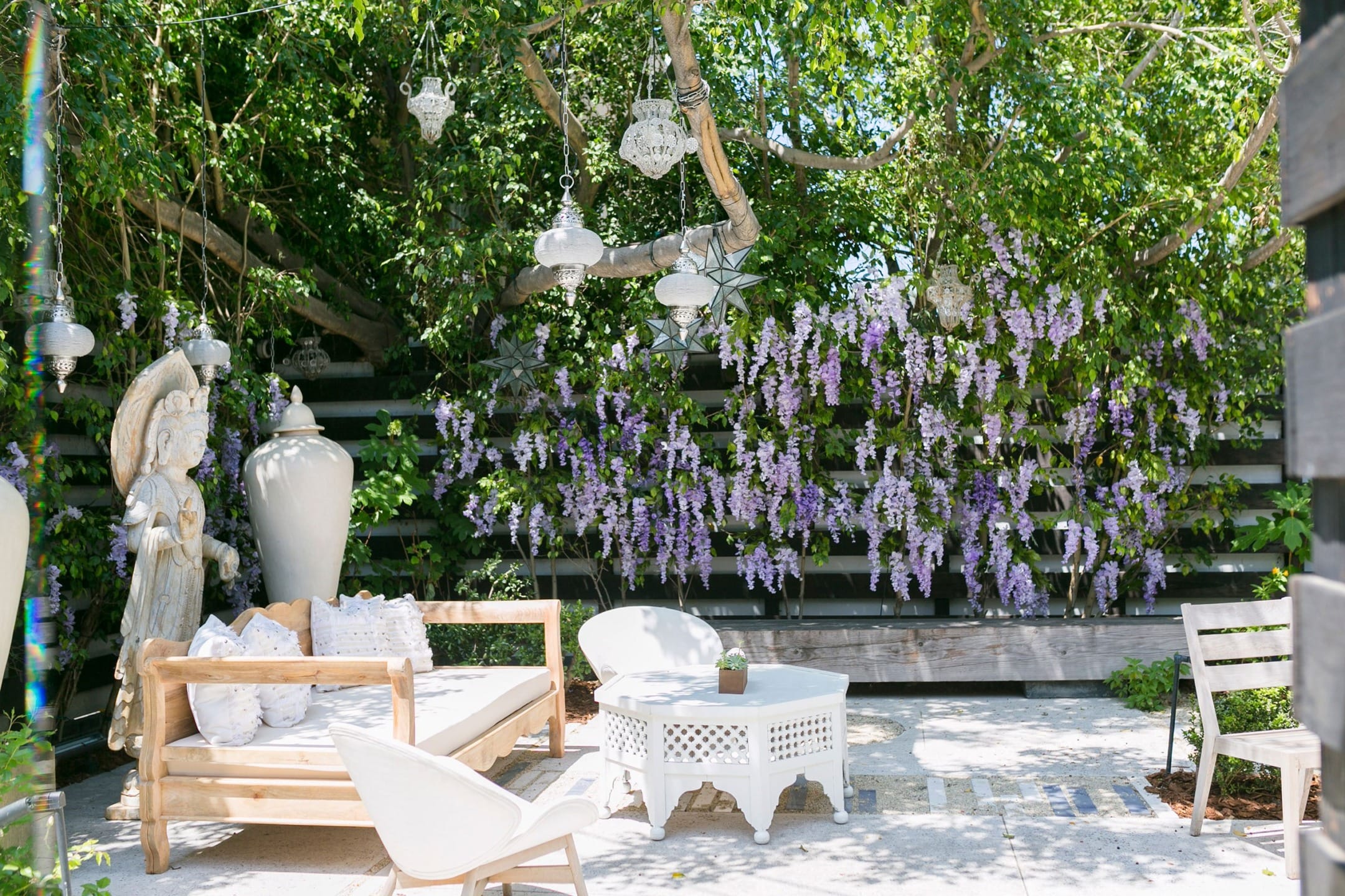 Outdoor garden seating area with wooden furniture, decorative lanterns, and blooming purple wisteria against a lush green backdrop.