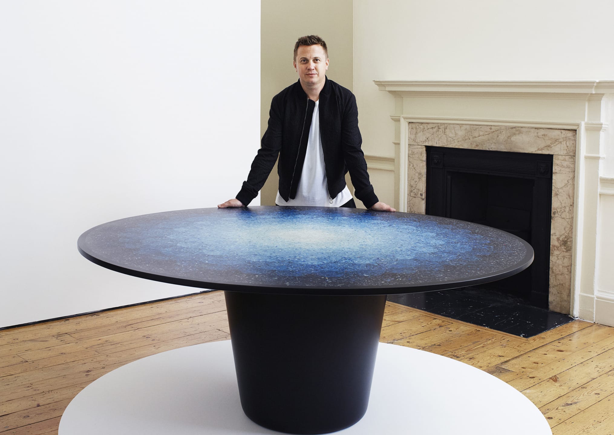 Man standing behind a large, round, artistic table with a blue pattern in a room with wooden floors and a fireplace.