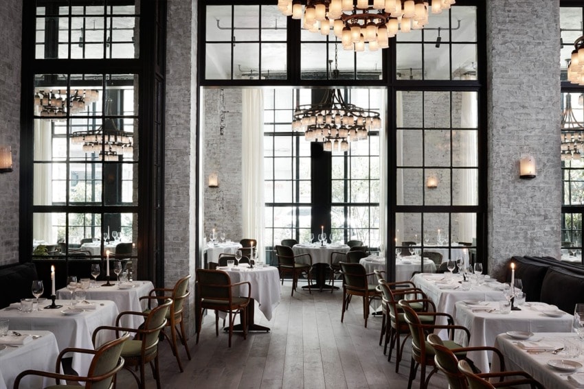 Elegant restaurant interior with white tablecloths, large windows, and chandelier lighting.